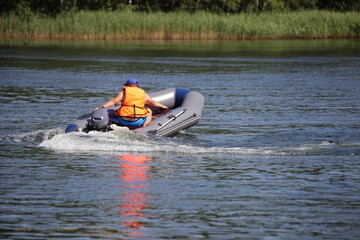 One man in orange lifejacket turns on inflatable motor boat with small outboard motor on grassy far shore background , active recreation on the water at Sunnny summer day