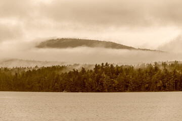 A tinted photograph of a misty morning on lake with low clouds covering the hills across the lake.