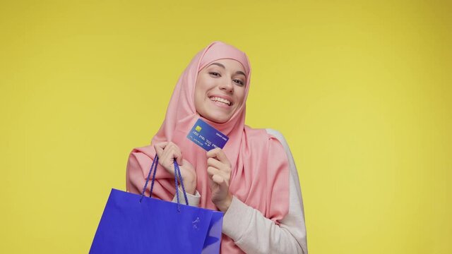 Beautiful young Muslim woman in hijab headscarf posing look at camera hold in hand credit bank card and hold in hands package bag with purchases after shopping isolated on yellow background in studio.