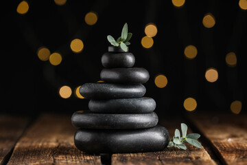 Obraz premium Stack of black spa stones with green branches on wooden table against blurred lights