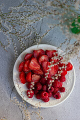 White plate with sliced strawberries and raspberries on a gray concrete background surrounded by small white flowers. Gentle spring mood, light snack