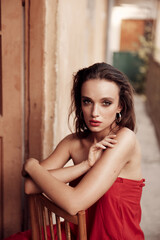 Naklejka premium Charismatic brunette in a long red dress with beautiful facial features. sitting on a chair on the balcony in a small Spanish street. Painted red lips and incredibly beautiful brown eyes