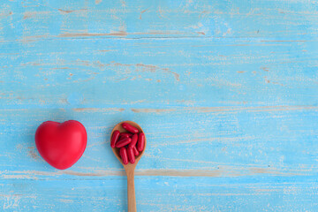 many red capsule medicines in wooden spoons for heart disease and healthcare with red heart on blue plank background