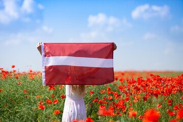 Blond girl holding flag of Latvia in the poppy field. Back view. Declaration of Independence Day....