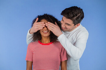 White man closes eyes of his beloved african american girlfriend before surprise her. interracial couple on blue studio background. Love, holiday, happiness concept.