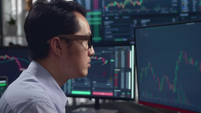 Sild View Of Asian Male Stock Market Broker Looking Analyzing Graphs On Multiple Computer Screens 
