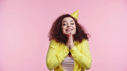 excited young woman in party cap with praying hands isolated on pink