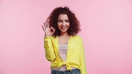 cheerful young and curly woman showing ok sign isolated on pink