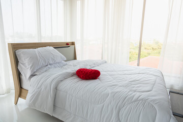 Interior white bedroom with curtains and white pillows, red heart shape pillow and duvet on wooden bed with window city view.