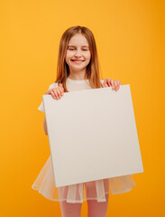 Cute pretty girl child holding white canvas and looking at the camera isolated on yellow background...