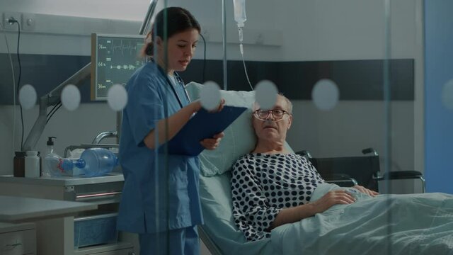 Caucasian Nurse Checking On Elder Patient In Hospital Ward At Medical Recovery Unit. Old Man With IV Drip Bag And Nasal Oxygen Tube Getting Help To Treat Heavy Health Problems And Illness