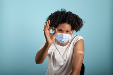 Young afro latino showing arm with coronavirus vaccine band-aid with ok gesture and protective mask on a blue background