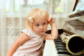 Fototapeta premium The cheerful little girl is leaning on the piano keys and pensively looks at the clarinet lying on the black piano keys.
