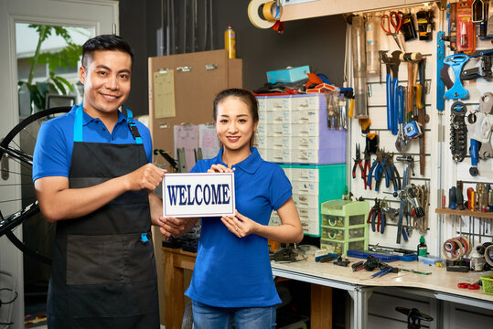 Portrait of young smiling bicycle workshop mechanics holding welcome sign when standing at wall with various tools