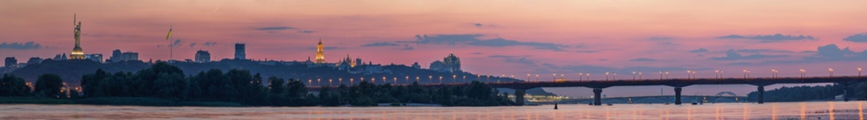  View of Kyiv Pechersk Lavra, Motherland Monument, Kyiv bridges and Dnieper river, after sunset a thin crescent moon is seen in the orange sky.