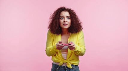 KYIV, UKRAINE - JUNE 30, 2021: worried young woman holding joystick and playing video game isolated on pink