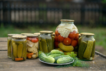 Glass jars with pickles and tomatoes on a wooden table in a rustic style. Plate with fresh cucumbers and dill.