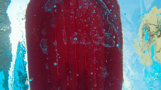 SLOW MOTION, UNDERWATER, LOW ANGLE, FLARE: From inside a pool a Underside red floating boat and the feet of a kid kicking while swimming. Boat Floating On Surface. Summer holidays vacation concept.