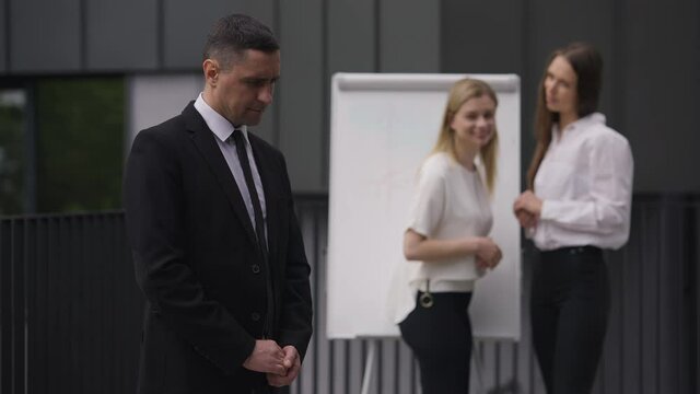 Portrait of shy male employee posing outdoors as blurred female colleagues gossiping laughing at background. Handsome Caucasian elegant man looking at camera as flirty women talking
