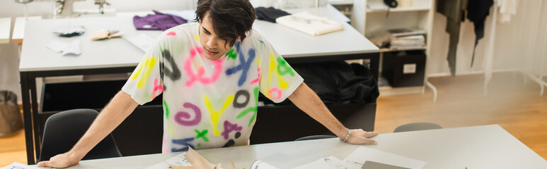Obraz na płótnie Canvas stylish designer standing near sewing patterns on desk in tailor shop, banner