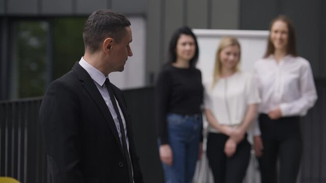 Portrait of confident man looking back at blurred colleagues crossing hands smiling. Satisfied Caucasian male CEO proud with team posing outdoors. Business and confidence concept