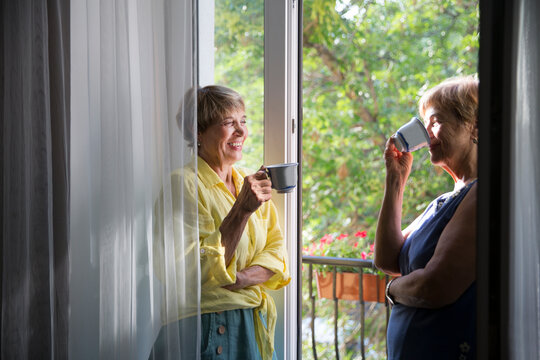 Portrait of happy senior woman friends standing on the balcony of the apartment and drink tea and talk
 - Powered by Adobe