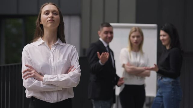 Portrait of confident arrogant woman crossing hands looking at camera ignoring colleagues mocking at background. Young beautiful Caucasian employee posing outdoors in slow motion