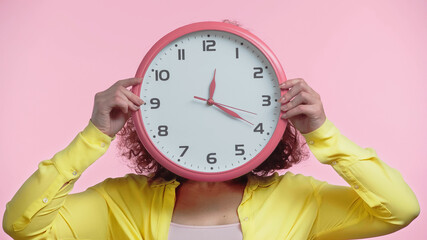 young woman covering face while holding clock isolated on pink