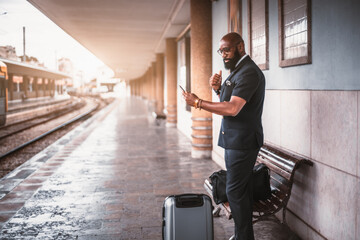 An elegant black businessman is standing with his baggage bags on an empty railrway platform of a railroad station depot and using his phone while waiting for a train to start his business trip