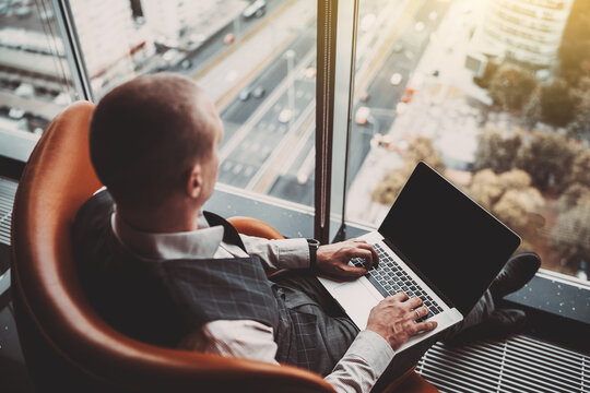 An elegant man entrepreneur is using his laptop while sitting in an armchair indoors of a luxury business skyscraper in front of the window with a highway outside; a businessman working on a netbook