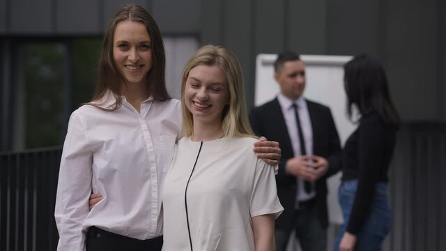 Happy young slim beautiful women looking at camera smiling hugging standing outdoors with colleagues talking at background. Positive Caucasian coworkers posing on meeting. Slow motion