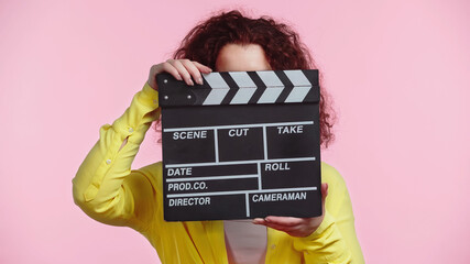 curly woman obscuring face with clapboard isolated on pink