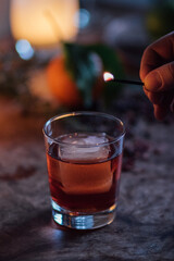 whiskey cocktail with large ice cube in rocks glass with hand holding a lit match 