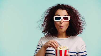 curly woman in 3d glasses eating popcorn while holding bucket isolated on blue