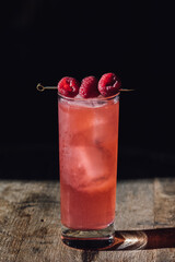 pink raspberry drink in tall Collins glass with ice and cocktail pick against black on wood barrel table 