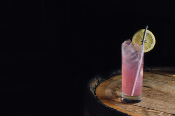 Pink lemonade in tall Collins glass with paper straw and lemon wheel on whiskey barrel against dark...