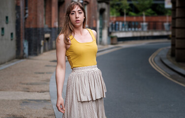 Fototapeta premium Young woman in a yellow t-shirt and white skirt posing in a narrow street.