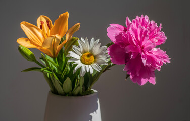 A bouquet of three different flowers in a vase on a grey background. Lily, chamomile and peony in a vase.