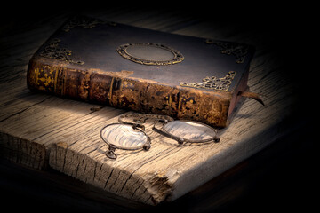 Libro con el lomo en piel desgastada con adornos de latón y sobe una tarima antigua de madera y con una gafas en un primer plano .
