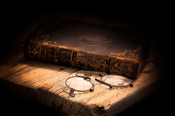 Imagen de un libro antiguo cerrado y con adornos metálicos en la portada . Situado sobre una tarima de madera y con unos binoculares en primer plano y con un fondo oscuro y cálido