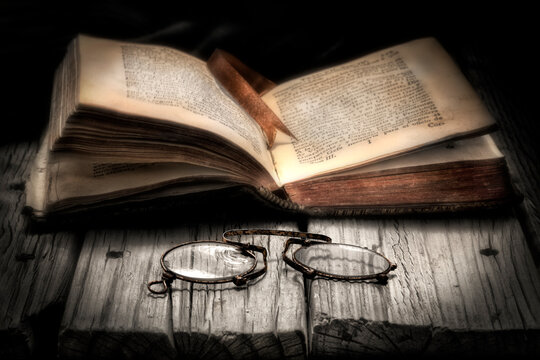 Imagen de un libro antiguo abierto sobre una mesa vieja de madera y con unos binoculares en primer plano. La imagen esta hecha en color y blanco y negro.