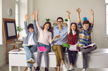 Education, successful completion of school year. Happy smiling teacher hugging overjoyed excited elementary pupils diverse group sitting on desk with raised hands up. Modern classroom interior