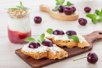 Sweet toasts with cream cheese, fresh ripe cherry and mint leaves decor on wooden board and miniature glass jar of aquafaba dessert with fruit jam and granola. Vegetarian snacks. Gluten free toasts.