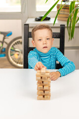 A cute seven-year-old child boy playing Jenga at home at a white wooden table against the background of a light window. Selective focus. Close-up. Portrait