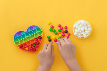 Use the popular pop it toy as a chocolate mold. Children is hands put colorful candies in a popit heart-shaped silicone mold on a yellow background. Step-by-step photo instructions. Step 2.