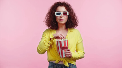 curly young woman in 3d glasses holding popcorn bucket isolated on pink