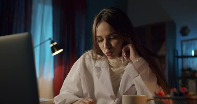 Young woman wearing medical uniform gown, female intern working at night with a computer in a medical office. Doctor nurse using laptop and video call, writing notes, overtime at the workplace.