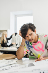 tired fashion designer sitting with closed eyes near sketches and colleague on blurred background