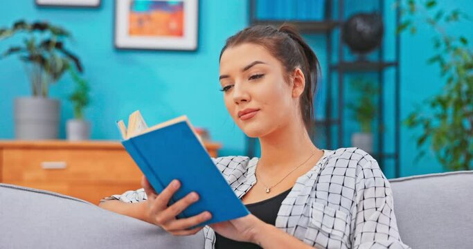 Young women with hair pinned up relaxes in homely attire while delving into book that is in hands, reads page with curiosity, analyzing meaning of novel, turning pages without stopping reading