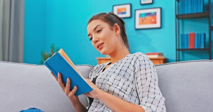 Pretty female with hair pinned up relaxes in homely attire while delving into book that is in hands, reads page with curiosity, analyzing meaning of novel, turning pages without stopping reading